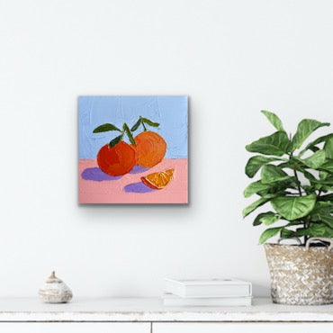 Canvas painting of oranges on a pink surface with a blue background, displayed on a wall above a white shelf.