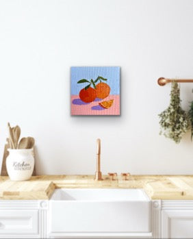 Framed artwork of apples and oranges on a kitchen wall with a sink and wooden cabinets.